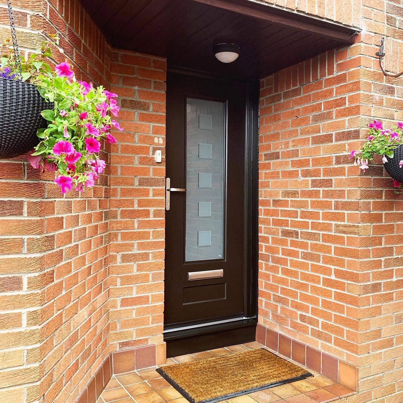 Dark entrance door with a tall glazed panel, brick porch and flowering hanging baskets